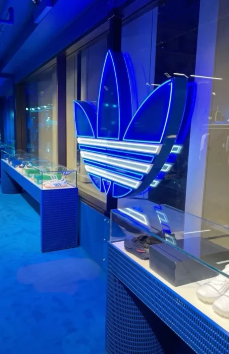 ntérieur d'une boutique Adidas Originals avec un logo tréfle lumineux en néon bleu et des sneakers exposées dans des vitrines en verre